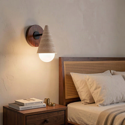 Travertine cone wall light lit above a bedside table beside a cane headboard