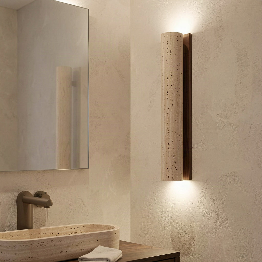 The travertine column wall light mounted beside a mirror in a stone-toned bathroom