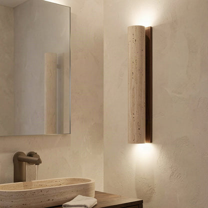 The travertine column wall light mounted beside a mirror in a stone-toned bathroom